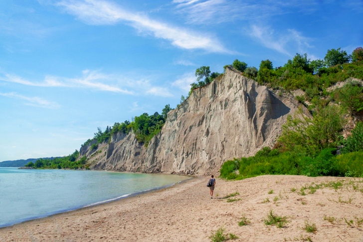 Woman Walks on Brown Seashore Near Cliff With Green Trees Under Blue and White Sky